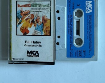 Może przedstawiać: Kasetę magnetofonową z albumem "Greatest Hits" Billa Haleya. Na okładce kolorowy obraz zespołu i tytuł albumu. Taśma oznaczona "MCA Records" i zawiera logo Dolby System.