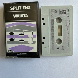Op de afbeelding: Een cassettebandje met een wit label en zwarte tekst. De tekst luidt "WAIATA Hard Act To Follow One Step Ahead. I Don't Wanna Dance. Iris. Wail. Clumsy SPLIT ENZ JMG". De cassetteband is gelabeld met "CAM 64848" en heeft een "AM Records" logo.