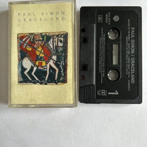 May include: A black cassette tape with the title "Paul Simon / Graceland" and the album's tracklist printed on the label. The cassette tape is in a clear plastic case with a colorful image of a man on a horse.