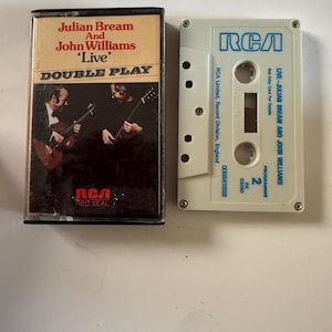 Puede incluir: Una cinta de casete y estuche vintage con el álbum "Double Play: Live" de Julian Bream y John Williams. El estuche tiene una foto de los músicos tocando la guitarra. La cinta es blanca con la marca RCA azul.