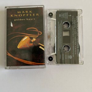 Könnte beinhalten: Ein Kassetten-Etui mit einer durchsichtigen Plastikhülle und einem schwarzen Kassettenband im Inneren. Die Vorderseite des Etuis zeigt ein Foto eines goldenen Herzens vor braunem Hintergrund. Der Text "MARK KNOPFLER - GOLDEN HEART" ist auf dem Etui aufgedruckt.
