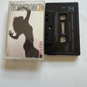 Puede incluir: Un casete Technotronic Pump Up The Jam y su caja de plástico transparente. La carátula muestra la silueta negra de una persona bailando sobre un fondo blanco con el texto Technotronic y pump up the jam. El casete negro tiene texto en blanco, incluyendo el número 2 y el título Pump Up The Jam.
