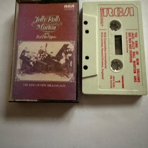 以下が含まれることがあります： 「Jelly Roll Morton and His Red Hot Peppers」をフィーチャーした、ヴィンテージのオーディオカセットテープとケース。カセットケースは、バンドの写真とテキストが入ったバーガンディ色です。カセットはオフホワイトで、赤いRCAのブランドが入っています。