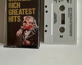 Może przedstawiać: Kasetę magnetofonową z albumem "Greatest Hits" Charliego Richa. Kaseta ma brązowe etui ze złotymi literami i zdjęciem Charliego Richa. Kaseta jest jasnoszara z tytułem albumu i tytułami piosenek.