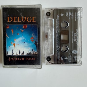 Puede incluir: Una cinta de cassette vintage con el álbum "DELUGE" de Jocelyn Pook. La portada muestra un cielo azul con formas y siluetas naranjas. La cinta está en una caja de plástico transparente.