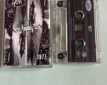 May include: A black cassette tape with the band name "Ash" and the album title "1977" printed on the front. The cassette tape is in a clear plastic case.