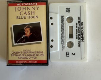 May include: A white cassette tape with a black label. The label says "Autograph Johnny Cash Blue Train" and features a photo of Johnny Cash. The tape is labeled "Ask 753" and "2".
