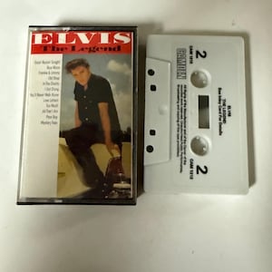 May include: A white cassette tape with a black label. The label has the text "ELVIS The Legend" and "CAM 1212" printed on it. The cassette tape is in a clear plastic case with a black border. The case has a picture of Elvis Presley on the front. The picture shows Elvis Presley standing in a field with a microphone in his hand. The text on the case reads "ELVIS The Legend" and lists the songs on the cassette tape: "Good Rockin' Tonight", "Blue Moon", "Frankie & Johnny", "Old Shep", "In the Ghetto", "I Got Strings", "Love Letters", "You'll Never Walk Alone", "Too Much", "All That I Am", "Poor Boy", "Mystery Train".