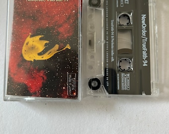 Puede incluir: Una cinta de casete con una etiqueta en blanco y negro que dice "NewOrder/TrueFaith-94". La cinta de casete está en una caja de plástico transparente. La cinta de casete es una grabación de música.