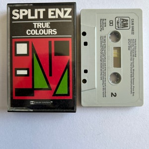 Może przedstawiać: Kaseta magnetofonowa z czarno-białą etykietą z napisem "SPLIT ENZ TRUE COLOURS" i czerwoną, zieloną i czarną grafiką. Kaseta jest oznaczona jako "CAM 64822" i zawiera tekst "TRUE COLOURS" oraz "SPLIT ENZ" na etykiecie.