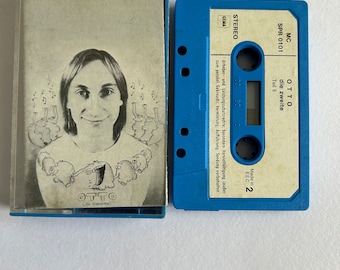 May include: A vintage audio cassette tape with a blue plastic case and a blue tape. The cassette case features a black and white illustration of a person with long hair and a cartoon design. The tape is labelled "OTTO die zweite".