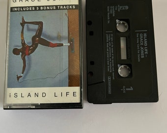 May include: A black cassette tape with a white label. The label has the text "Island Life - Grace Jones" and "See Inlay Card For Details". The cassette tape is in a black plastic case.
