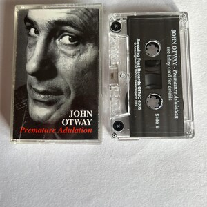 May include: A black and white cassette tape with a photo of John Otway on the front. The tape is titled "Premature Adulation" and is a release from Amazing Feet Records.