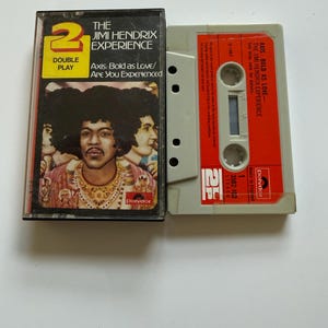 Könnte beinhalten: Eine Kassette und ihre Kunststoffhülle mit The Jimi Hendrix Experience, Axis: Bold as Love/Are You Experienced. Das Cover zeigt ein Porträt von Jimi Hendrix, während die graue Kassette ein leuchtend orangefarbenes Etikett mit schwarzem Text hat.