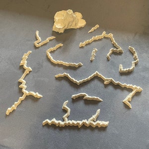 May include: A white 3D printed terrain set for tabletop gaming. The set includes a mountain and a variety of winding paths.