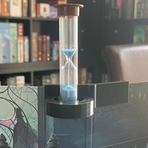 May include: A blue hourglass with a brown top is mounted on a black stand. The hourglass is attached to a black board with a decorative design.