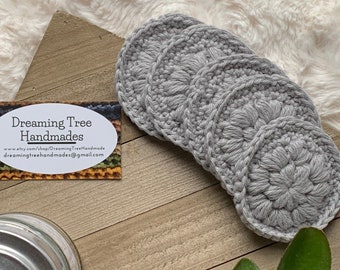 Peut inclure: Quatre sous-verres en crochet gris avec un motif floral en relief. Les sous-verres sont posés sur une surface en bois avec une carte de visite qui indique "Dreaming Tree Handmades" avec un site web et une adresse e-mail.