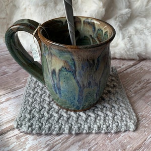 May include: A green and brown ceramic mug with a gray crocheted coaster. The mug has a handle and a spoon inside.