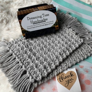 May include: Two gray crocheted coasters with fringe edges. The coasters are stacked on top of each other. A business card for "Dreaming Tree Handmades" is in the background. A "Thank You" sticker is on a pink and white patterned surface.