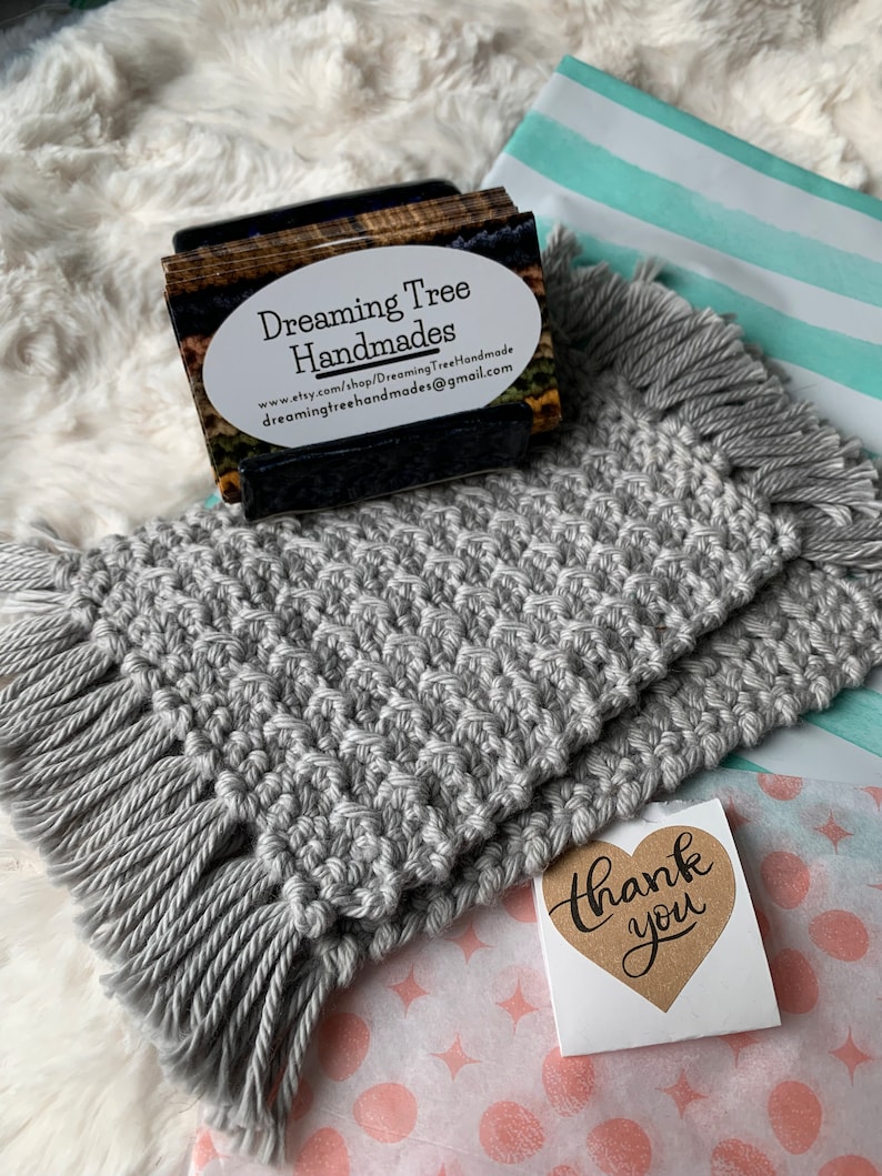 May include: Two gray crocheted coasters with fringe edges. The coasters are stacked on top of each other. A business card for "Dreaming Tree Handmades" is in the background. A "Thank You" sticker is on a pink and white patterned surface.