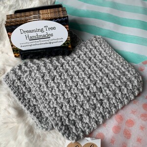 May include: Gray crocheted pot holder with a thank you tag. The tag has a heart shape with the words "thank you" written in black cursive. The pot holder is on a white fluffy surface.