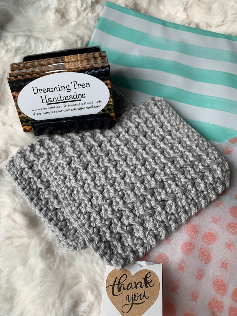 May include: Gray crocheted pot holder with a thank you tag. The tag has a heart shape with the words "thank you" written in black cursive. The pot holder is on a white fluffy surface.