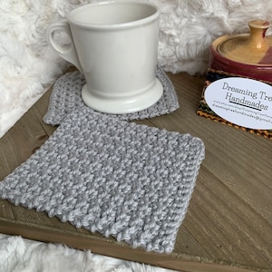 May include: Two gray knitted coasters with a white ceramic mug on top of one of the coasters. The coasters are on a wooden surface. A business card with the text "Dreaming Tree Handmades" is on the surface. The business card includes a website address and email address.