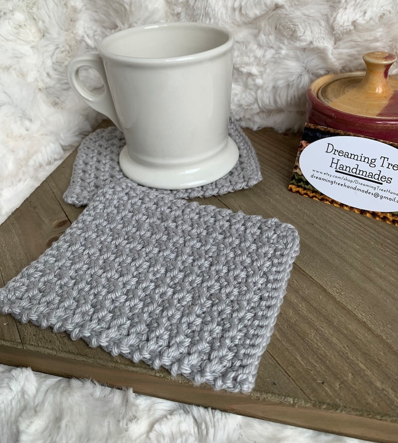 May include: Two gray knitted coasters with a white ceramic mug on top of one of the coasters. The coasters are on a wooden surface. A business card with the text "Dreaming Tree Handmades" is on the surface. The business card includes a website address and email address.