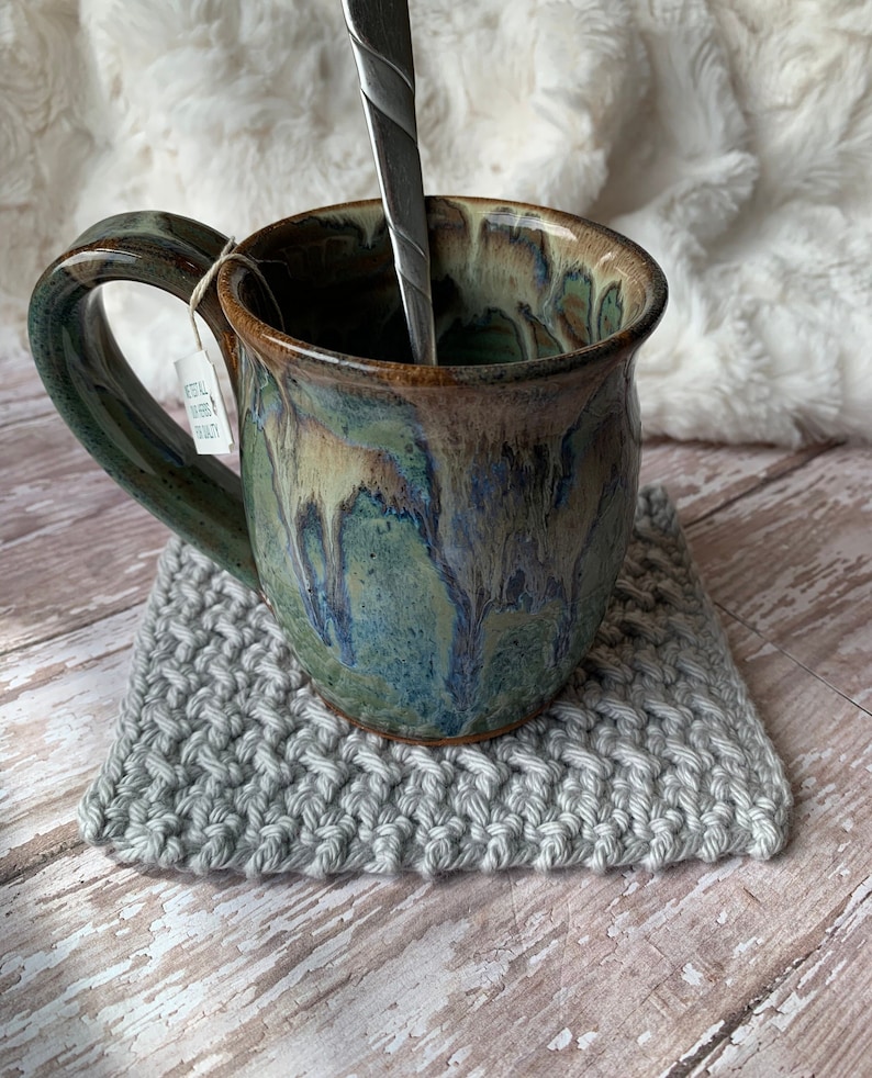 May include: A green and brown ceramic mug with a gray crocheted coaster. The mug has a handle and a spoon inside.