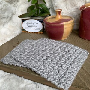 May include: A gray crocheted coaster with a woven pattern. The coaster is on a wooden surface with a small ceramic jar and a business card that reads "Dreaming Tree Handmades".