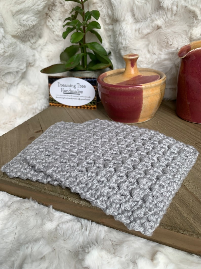 May include: A gray crocheted coaster with a woven pattern. The coaster is on a wooden surface with a small ceramic jar and a business card that reads "Dreaming Tree Handmades".