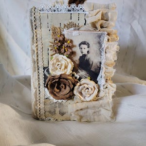 May include: A handmade journal with a vintage aesthetic. The cover features a sepia-toned portrait, fabric ruffles, lace trim, and paper roses in cream and brown. A small brown feather accents the top.