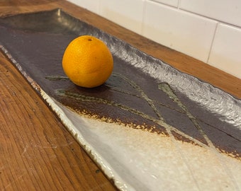 Könnte beinhalten: Ein rechteckiger Keramik-Servierteller mit brauner und weißer Glasur, akzentuiert mit grünen Pinselstrichen. Eine orangefarbene Zitrusfrucht liegt auf dem Teller. Der Teller steht auf einer Holzoberfläche.