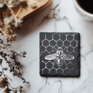 May include: A square, black slate coaster featuring a detailed illustration of a bee. The bee is centered on a honeycomb pattern. The coaster is on a marble surface, with a wooden tray and a cup of coffee in the background.