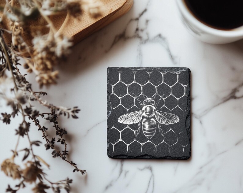 May include: A square, black slate coaster featuring a detailed illustration of a bee. The bee is centered on a honeycomb pattern. The coaster is on a marble surface, with a wooden tray and a cup of coffee in the background.