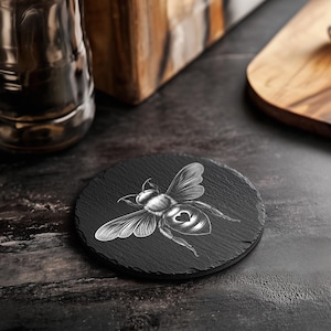 May include: A round, black slate coaster with a detailed white illustration of a bee. The bee has a heart-shaped design on its abdomen. The coaster is on a dark surface, with a glass and wooden cutting board in the background.
