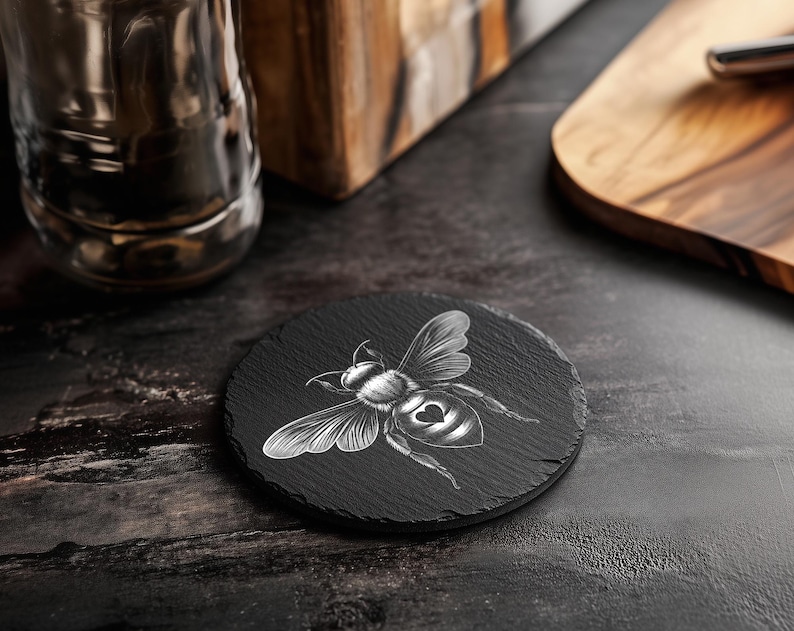 May include: A round, black slate coaster with a detailed white illustration of a bee. The bee has a heart-shaped design on its abdomen. The coaster is on a dark surface, with a glass and wooden cutting board in the background.