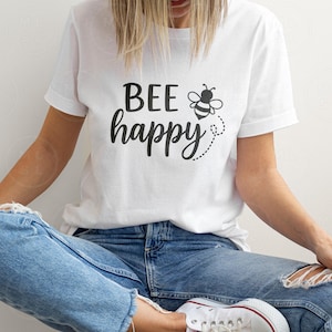 May include: White t-shirt with the phrase "BEE happy" in black script, accompanied by a bee graphic. The shirt is worn with blue jeans. The overall theme is positivity and nature.