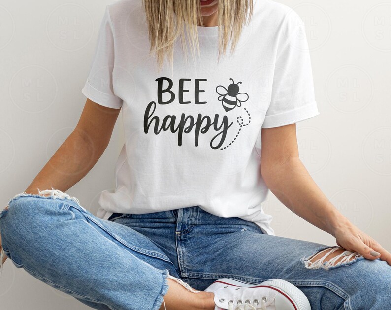 May include: White t-shirt with the phrase "BEE happy" in black script, accompanied by a bee graphic. The shirt is worn with blue jeans. The overall theme is positivity and nature.