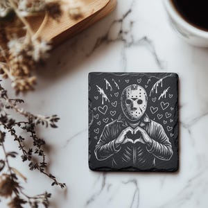 May include: Black square coaster featuring a white illustration of a hockey mask-wearing figure making a heart shape with their hands. White hearts and drips surround the figure. The coaster is on a marble surface.