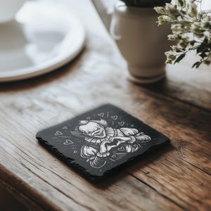 May include: A black square coaster with a white illustration of a clown with hearts. The coaster is made of a dark, textured material and sits on a wooden surface. The clown has a ruffled collar and is surrounded by small heart shapes.