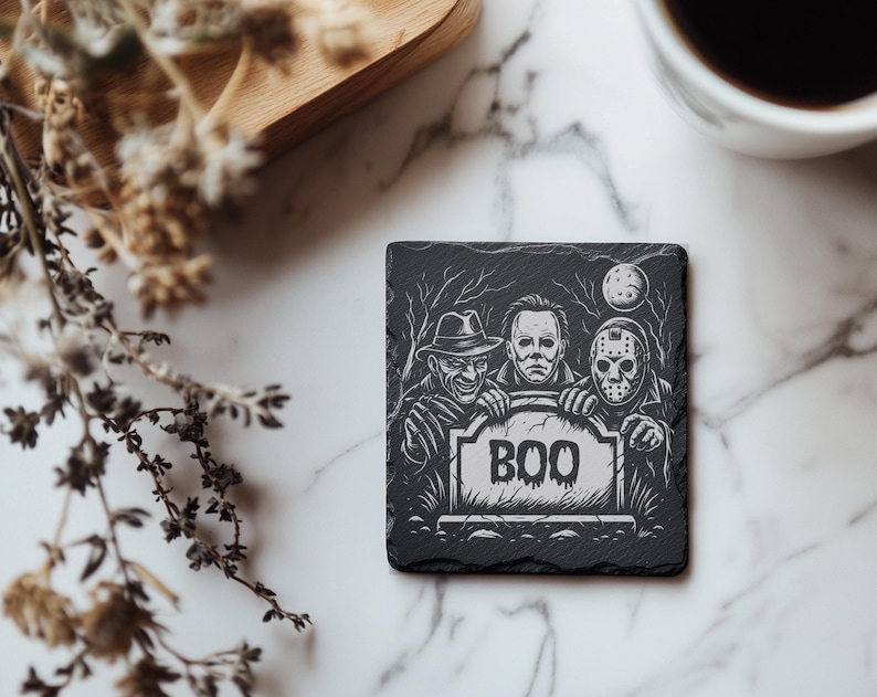 May include: Black square coaster with a Halloween theme. The coaster features three horror movie characters holding a tombstone that says "BOO". The design is in black and white.