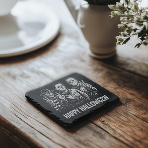 May include: A black square slate coaster with a Halloween theme. It features illustrations of Freddy Krueger, Michael Myers, and Jason Voorhees, with the words "HAPPY HALLOWEEN" below the images. The coaster is on a wooden surface.