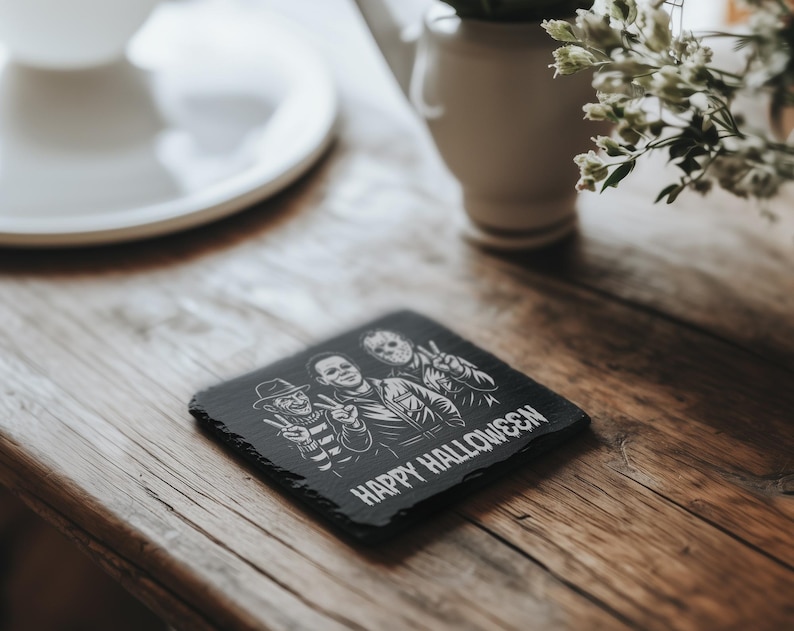 May include: A black square slate coaster with a Halloween theme. It features illustrations of Freddy Krueger, Michael Myers, and Jason Voorhees, with the words "HAPPY HALLOWEEN" below the images. The coaster is on a wooden surface.