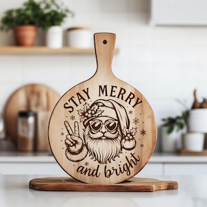 May include: A wooden cutting board with a handle, featuring a laser-etched design of Santa Claus wearing sunglasses and giving a peace sign. The text "STAY MERRY and bright" is also etched on the board. The board is round with a handle.