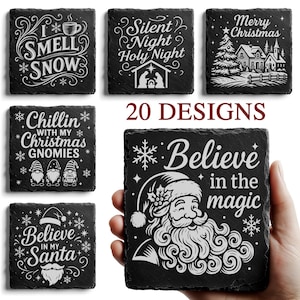 May include: A collection of black slate coasters with white engraved holiday designs. The coasters feature festive phrases like "I Smell Snow," "Merry Christmas," and "Believe in Santa." One coaster is held in a hand, displaying Santa Claus.