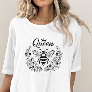 May include: White t-shirt featuring a black graphic of a bee inside a floral wreath, with the word "Queen" above. The design includes a small crown. The shirt has a round neckline and short sleeves.