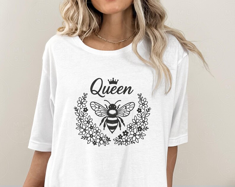 May include: White t-shirt featuring a black graphic of a bee inside a floral wreath, with the word "Queen" above. The design includes a small crown. The shirt has a round neckline and short sleeves.