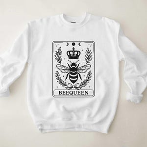 May include: White sweatshirt with a black graphic of a bee wearing a crown, surrounded by leaves and moons. The word "BEEQUEEN" is printed at the bottom of the design. The sweatshirt has long sleeves and a crew neck.