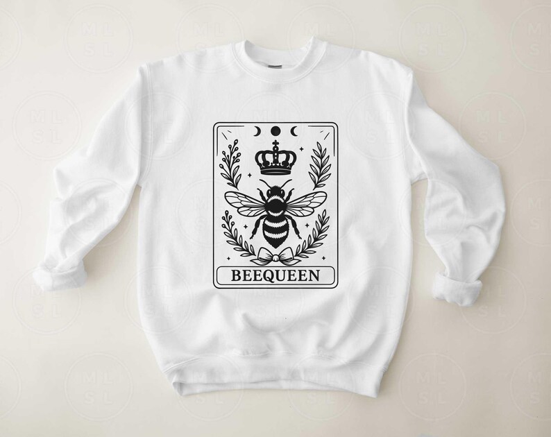 May include: White sweatshirt with a black graphic of a bee wearing a crown, surrounded by leaves and moons. The word "BEEQUEEN" is printed at the bottom of the design. The sweatshirt has long sleeves and a crew neck.